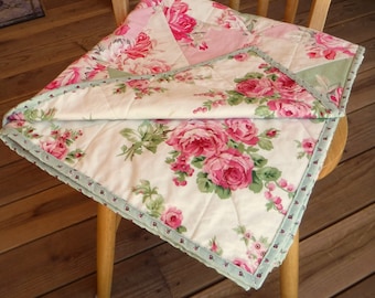 May include: A white quilt with a floral pattern featuring pink roses and green leaves. The quilt has a light green trim and is folded in half on a wooden chair.