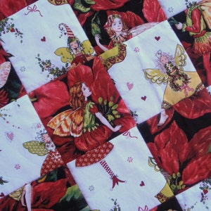 May include: A patchwork quilt with a Christmas theme featuring red poinsettia flowers and fairies with wings. The quilt is made of white fabric with red and green accents.