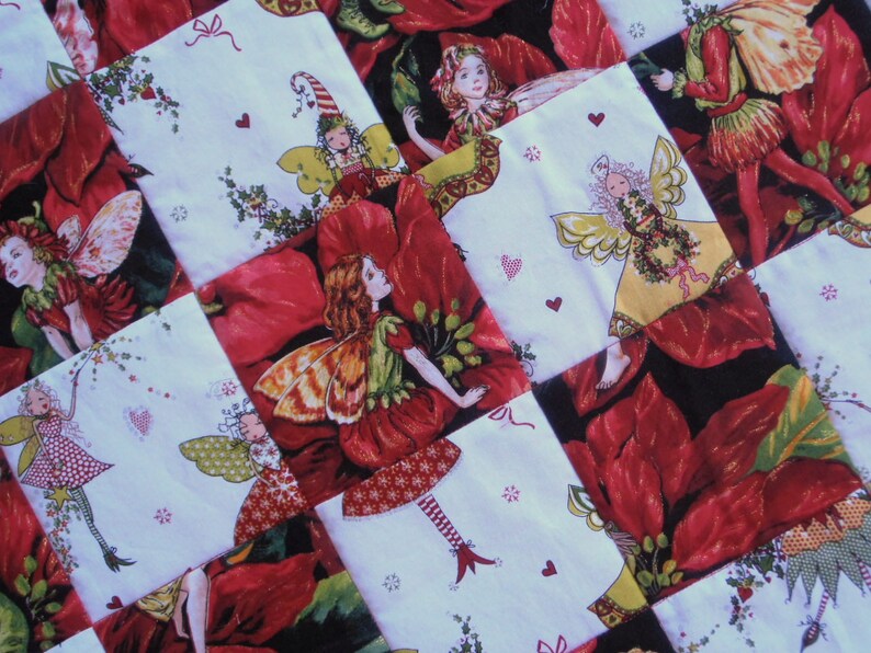 May include: A patchwork quilt with a Christmas theme featuring red poinsettia flowers and fairies with wings. The quilt is made of white fabric with red and green accents.