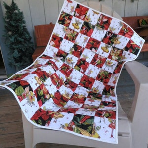 May include: A white and red patchwork quilt with a Christmas theme. The quilt features a repeating pattern of red poinsettias and fairies with wings. The quilt is folded and displayed on a white chair.