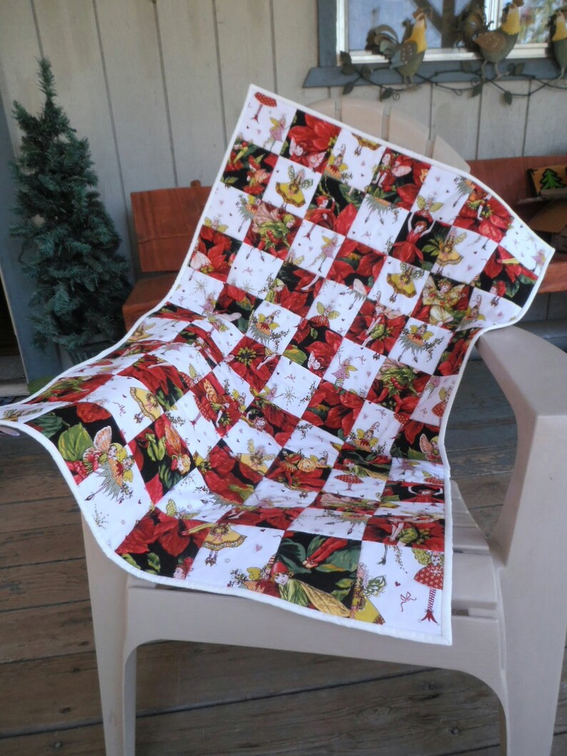 May include: A white and red patchwork quilt with a Christmas theme. The quilt features a repeating pattern of red poinsettias and fairies with wings. The quilt is folded and displayed on a white chair.