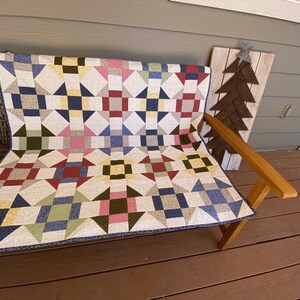 May include: A rectangular quilt featuring a repeating block pattern of geometric shapes in various colors including navy blue, red, yellow, pink, and olive green, set against a cream-colored background with a dark blue patterned border.
