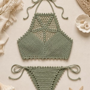 Pode incluir: Um conjunto de biquíni de crochê na cor verde sálvia, composto por um top estilo frente única com um decote de renda vazada intrincado e uma parte de baixo combinando com amarrações laterais. Ambas as peças têm um delicado acabamento em picot.