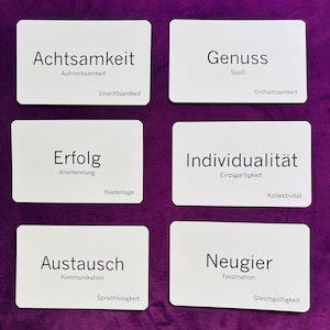 Op de afbeelding: Zes witte rechthoekige kaarten met Duitse woorden en gerelateerde termen liggen op een paarse ondergrond. De kaarten tonen Achtsamkeit, Genuss, Erfolg, Individualität, Austausch en Neugier. Geschikt voor een spel of als leermiddel.