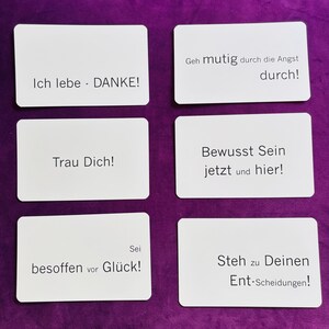 Könnte beinhalten: Sechs weiße rechteckige Karten mit schwarzem Text auf lila Hintergrund. Die Karten enthalten verschiedene deutsche Sätze, darunter "Ich lebe - DANKE!", "Geh mutig durch die Angst durch!" und "Trau Dich!"