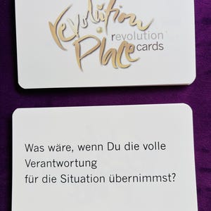 May include: Two white rectangular cards. The top card has gold script that reads "Revolution Place cards". The bottom card has German text: "Was wäre, wenn Du die volle Verantwortung für die Situation übernimmst?"