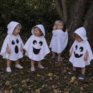 May include: Four white hooded ponchos featuring black ghost face graphics. Three ponchos display distinct facial expressions with large eyes and open mouths, while one poncho is plain white with a hood. The garments are worn by four small children.
