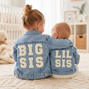 Könnte beinhalten: Zwei hellblaue Jeansjacken mit weißer, strukturierter Beschriftung im College-Stil auf der Rückseite. Die größere Jacke zeigt den Schriftzug BIG SIS, die kleinere Jacke den Schriftzug LIL SIS. Beide Jacken haben klassische Kragen und Knöpfe.