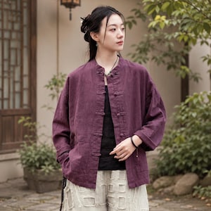 May include: A deep purple long-sleeved jacket featuring a mandarin collar and five fabric-covered toggle closures down the front. The jacket is crafted from a textured fabric and includes two front pockets, worn over a black top and light-colored pants.