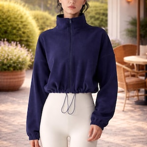 May include: A woman wearing a navy blue fleece zip-up jacket with a high collar and white leggings. The jacket has long sleeves and a drawstring at the waist. The leggings are high-waisted. The background is a blurred outdoor scene.