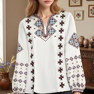 May include: A white long-sleeved blouse featuring a V-neckline and intricate geometric embroidery in shades of dark red, light blue, and black. The pattern is displayed along the neckline, down the front, and across the sleeves.