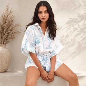 May include: White shirt and shorts co-ord set with a light blue illustrated design. The set includes a collared shirt with short sleeves and matching shorts. The design features drawings of flowers, cocktails, and other decorative elements.