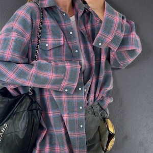 Può includere: Una camicia a quadri grigia, rosa e blu con due tasche sul petto e bottoni bianchi. La camicia è indossata aperta sopra una maglietta bianca e abbinata a jeans scuri. È visibile una borsa tote nera con tracolla a catena.