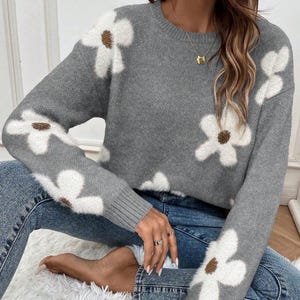 May include: A grey knitted jumper with large white daisy patterns and brown centres. The long-sleeved jumper has a crew neck and ribbed cuffs. The model is wearing blue jeans.
