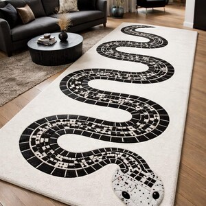 Puede incluir: Una alfombra rectangular grande con un diseño de serpiente. La alfombra presenta un fondo crema con un motivo de serpiente en blanco y negro que serpentea por la superficie. La cabeza de la serpiente está detallada con un patrón texturizado y una lengua rosa.