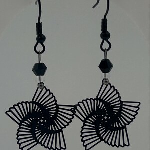 Peut inclure: Boucles d'oreilles noires en forme d'étoile avec un design ajouré unique. Chaque boucle d'oreille présente un motif en spirale à l'intérieur de l'étoile, suspendue à des crochets noirs avec de petites perles noires et une perle noire facettée. Un accessoire élégant.