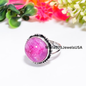 May include: A silver ring featuring a round, polished pink stone with a subtle grid-like pattern. The metal setting has a decorative beaded edge. The text Maria925JewelsUSA is printed on the right side of the ring band and stone setting.
