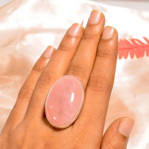 Peut inclure: Une bague en quartz rose ovale avec un anneau en argent. La pierre précieuse rose polie est sertie dans un design simple et élégant. La bague est présentée sur une main.