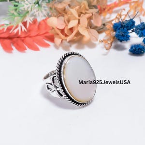May include: A silver ring featuring a large, smooth, oval-shaped white stone set in a twisted rope-style border. The band displays an openwork design with cutouts. The text Maria925JewelsUSA is visible next to the ring on the white surface.