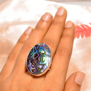 May include: Oval-shaped silver ring featuring a vibrant abalone shell. The shell displays iridescent blues, greens, and purples. The ring is set in a silver band, showcasing the natural beauty of the gemstone. A statement piece for jewelry lovers.