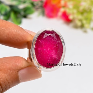 May include: A silver ring featuring a large, oval-shaped, faceted hot pink gemstone set in a smooth metal bezel. The text Maria925JewelsUSA is printed in black on the lower right side of the stone. The ring has a polished silver band.