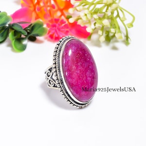 May include: A silver ring featuring a large, oval-shaped, vibrant magenta gemstone with a textured surface. The setting includes a beaded border and intricate scrollwork along the sides of the band. The text Maria925JewelsUSA is visible on the side.