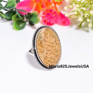 Pode incluir: Um anel de formato oval com uma pedra de coral fossilizado bege com um padrão de favo de mel distinto, inserido numa banda de prata polida com uma borda de corda torcida. O texto Maria925JewelsUSA é visível no canto inferior direito da imagem.