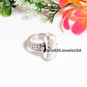 Può includere: Un anello in argento sterling con una perla bianca allungata e irregolare incastonata in una lunetta liscia. La fascia presenta un motivo dettagliato a foglie ripetute sull'esterno. Il testo Maria925JewelsUSA è visibile a destra.