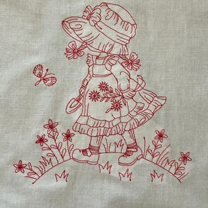 May include: Red embroidered design of a girl in a dress and hat walking in a field of flowers. The girl is looking up at a butterfly.