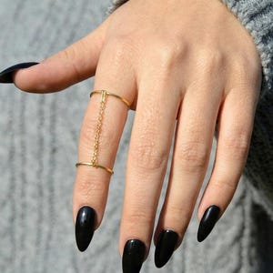 May include: A gold-toned finger ring featuring two thin bands connected by a delicate vertical chain. The upper band sits above the knuckle, while the lower band rests at the base of the finger, creating a linked design that spans the middle digit.