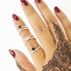 May include: A hand displays several silver rings. Two rings feature a small black stone, while another has a dragonfly design. The nails are painted a deep red, and the hand is adorned with intricate henna art.