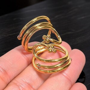 May include: A gold-toned metal ring featuring a multi-layered spiral design that wraps around the finger. The ends of the wire are finished with small, clustered spherical beads, creating a distinct decorative accent on this coiled jewelry piece.
