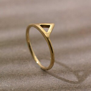 May include: A gold ring with a triangular design. The ring has a slender band that curves up to form the triangle. The ring is set against a neutral background.