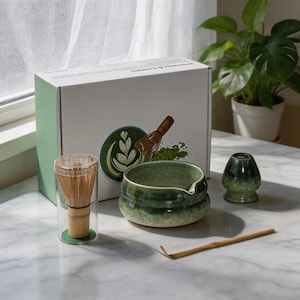 May include: A matcha tea set including a bamboo whisk in a clear cylindrical case, a green ceramic bowl with a pouring spout, a green ceramic whisk holder, and a bamboo scoop. The set is presented with a white box featuring a green logo.