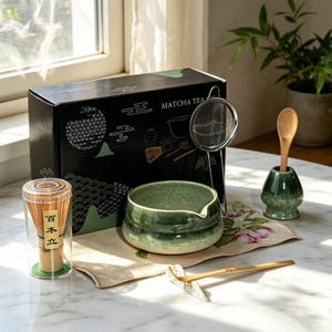 May include: A matcha tea set including a black box with white graphics, a green ceramic bowl, a bamboo whisk in a clear case, a metal strainer, a bamboo spoon in a green ceramic holder, a bamboo scoop on a rest, and a floral patterned cloth.