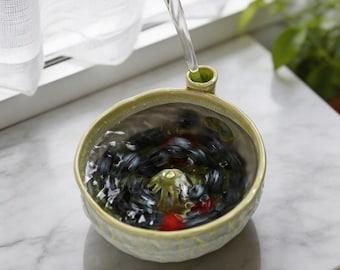 May include: A light green ceramic berry colander bowl with a small spout and a central drainage feature. The bowl is filled with water and dark blue berries, with a stream of water pouring into the top, creating a clear view of the fruit inside.