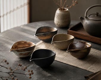 May include: A collection of six ceramic tea bowls in various earth-tone colors, including black, brown, and beige. Each bowl has a spout and is arranged on a dark gray surface with a neutral-colored runner. A teapot and vase are also visible.
