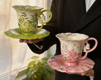 May include: Two ceramic teacups with matching saucers, one in lime green with yellow floral patterns and one in soft pink with pink floral patterns. Both cups feature scalloped rims and handles shaped like white lily of the valley flowers.