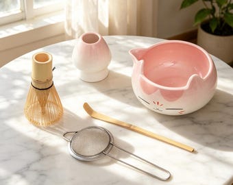 May include: A matcha tea set with a bamboo whisk, a pink cat-shaped bowl, a small pink vase, a bamboo scoop, and a stainless steel strainer. The items are arranged on a white marble table.