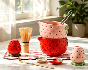 May include: A collection of strawberry-themed kitchenware, including two ceramic bowls, a whisk, a spoon, and small strawberry-shaped containers. The bowls are red and pink with dark red dots. The whisk is made of bamboo.