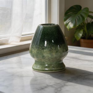 May include: A ceramic matcha whisk holder featuring a glossy finish with a gradient color scheme that transitions from a deep forest green at the top to a lighter, mottled sage green at the base, with a smooth, rounded, and tapered silhouette.