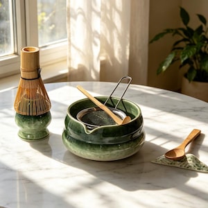 May include: A matcha tea set featuring a green ceramic whisk holder with a bamboo whisk, a green ceramic bowl containing a metal mesh strainer and a bamboo scoop, and a small green ceramic spoon rest with a wooden spoon.