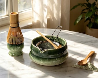 May include: A matcha tea set featuring a green ceramic whisk holder with a bamboo whisk, a green ceramic bowl containing a metal mesh strainer and a bamboo scoop, and a small green ceramic spoon rest with a wooden spoon.