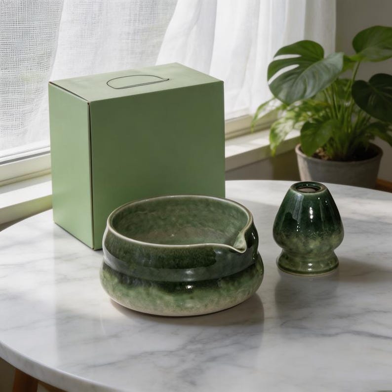 May include: A matcha bowl with a pouring spout and a matching whisk holder, both featuring a glossy, mottled green glaze. A light green cardboard box with a handle is positioned behind the ceramic items, which have a smooth, rounded shape.