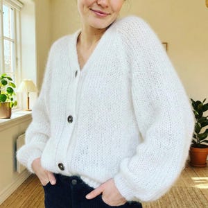 May include: A white, fluffy cardigan sweater with a V-neck and three dark buttons. The sweater has long sleeves and a relaxed fit. The person is wearing dark blue pants. The background includes a window, a lamp, and a potted plant.