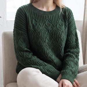 May include: A dark forest green long-sleeve sweater featuring a prominent knit leaf pattern across the front and sleeves. The garment has a ribbed crew neckline, ribbed cuffs, and a ribbed hem, showcasing a detailed textured stitch design.