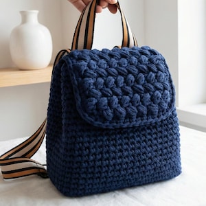 May include: A navy blue crochet backpack with a textured, raised pattern on the flap. The bag has striped straps in beige and brown. The bag is made of a soft, thick yarn.