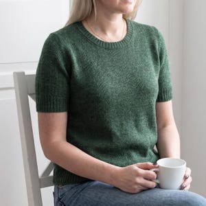 May include: A short-sleeved, forest green sweater with a crew neck. The sweater is made of a knit fabric and is worn with blue jeans. A white mug is held in the hands. The person is sitting on a white chair.