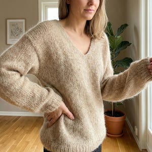 May include: A beige V-neck jumper with long sleeves. The jumper has a relaxed fit and is made from a soft, textured knit. The neutral colour makes it a versatile piece for any wardrobe.