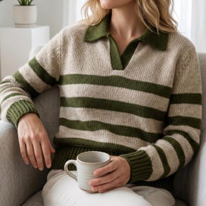 May include: A beige and green striped sweater with a collared neckline. The sweater has green stripes on the sleeves and body, with a green collar. A person is holding a ceramic mug. The sweater is made of a knitted material.